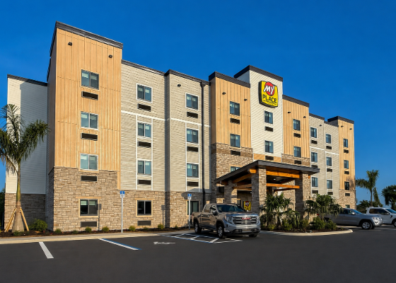 Exterior view of a My Place hotel with beige and stone façade, a covered entrance, and parked cars under a blue sky.