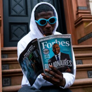 Person in a white hoodie wearing blue sunglasses reads a Forbes magazine outdoors on steps.