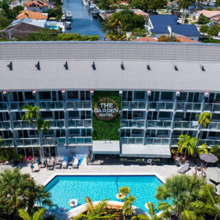 Aerial view of The Garden Hotel with a rectangular pool, palm trees, lounge chairs, and a canal in the background.