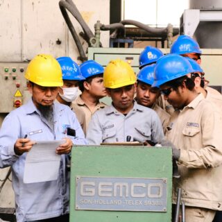 Group of factory workers wearing yellow and blue hard hats gathered around a green GEMCO machine, reviewing a sheet of paper.