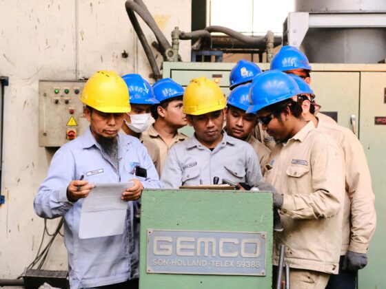 Group of factory workers wearing yellow and blue hard hats gathered around a green GEMCO machine, reviewing a sheet of paper.