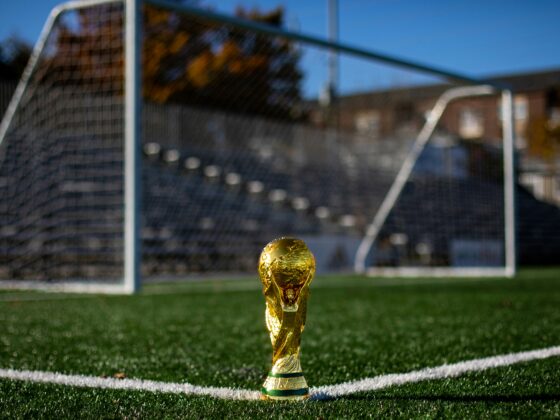 Golden World Cup trophy placed on a grassy soccer field with a blurred goal in the background.
