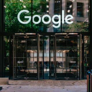 Glass entrance with a large illuminated Google logo on the glass facade, reflecting trees and the surrounding building.
