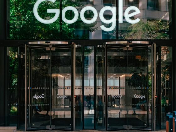 Glass entrance with a large illuminated Google logo on the glass facade, reflecting trees and the surrounding building.