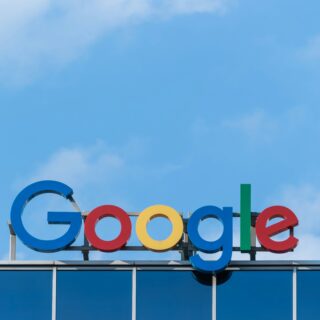 Google wordmark with multicolored letters mounted on a building under a blue sky.
