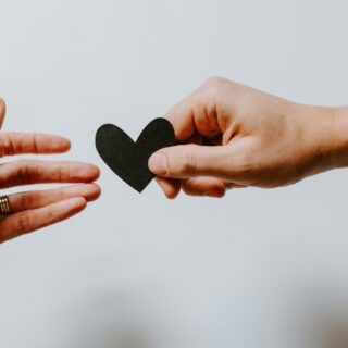 Two hands pass a black heart cutout between them against a pale background (love/connection symbol).