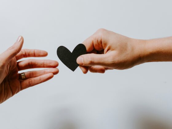 Two hands pass a black heart cutout between them against a pale background (love/connection symbol).