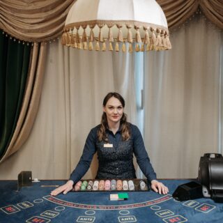Female casino dealer standing behind a blackjack table with stacked chips and cards, gold curtains in the background.