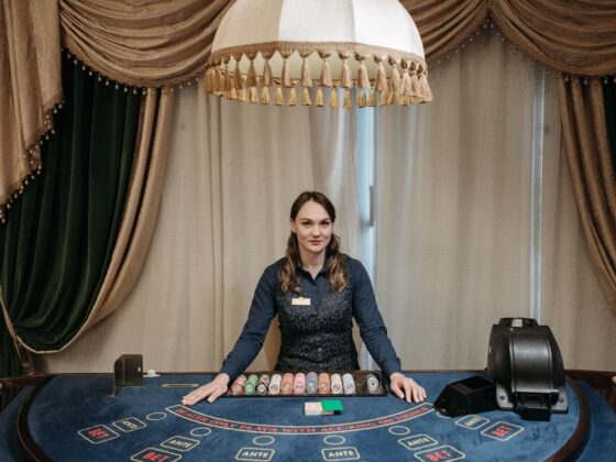 Female casino dealer standing behind a blackjack table with stacked chips and cards, gold curtains in the background.