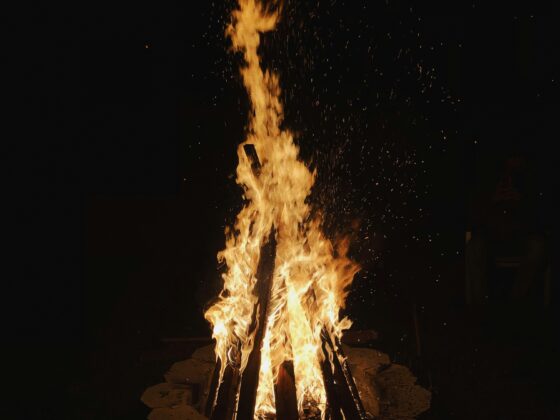 Night scene of a large bonfire with tall orange flames and flying embers against a dark sky, burning wooden logs in a circle outdoors.