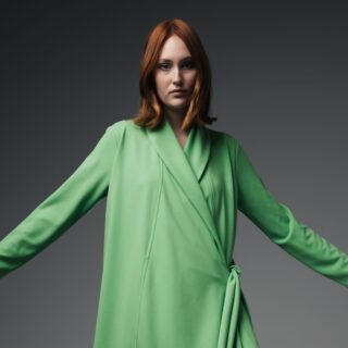Woman with short red hair wearing a lime-green wrap coat, arms extended against a dark gray gradient background.