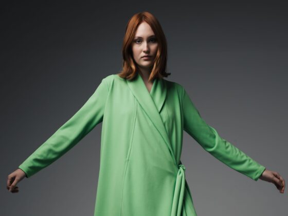 Woman with short red hair wearing a lime-green wrap coat, arms extended against a dark gray gradient background.