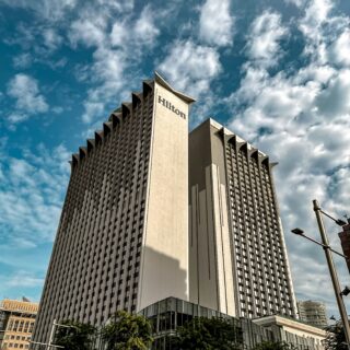 Tall Hilton hotel tower with stepped architectural features against a blue sky with scattered clouds.