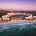 Aerial view of a large white beachfront resort with palm trees along a sandy shore at sunset.