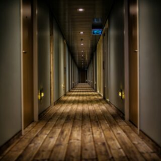 Dimly lit hotel hallway with closed doors on both sides and warm wooden floor, fading into distance at the end.