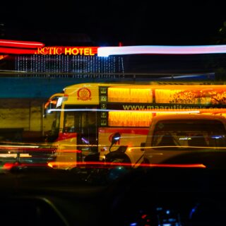 Night city street at night with long exposure light trails; a yellow tour bus in the foreground and a red Arctic Hotel sign in the background, blue string lights hanging across the scene.