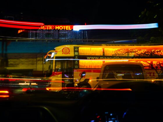 Night city street at night with long exposure light trails; a yellow tour bus in the foreground and a red Arctic Hotel sign in the background, blue string lights hanging across the scene.