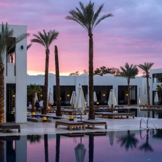 Resort pool at dusk with palm trees, white buildings, and pink-purple sunset skyc?