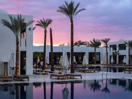 Resort pool at dusk with palm trees, white buildings, and pink-purple sunset skyc?