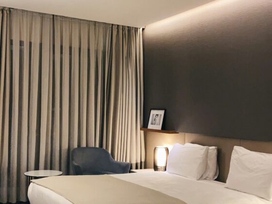 Hotel bedroom with a large bed, beige linens, and a gray accent wall; curtains, a blue chair, round side table, and bedside lamp.