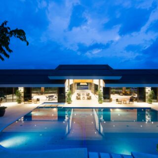 Evening view of a modern hotel pool area with lounge chairs around a rectangular pool and a glass-walled lobby behind, under a blue dusk sky.
