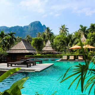 Turquoise poolside at a tropical resort with thatched villas, palm trees, and orange umbrellas, set against green hills and blue sky.