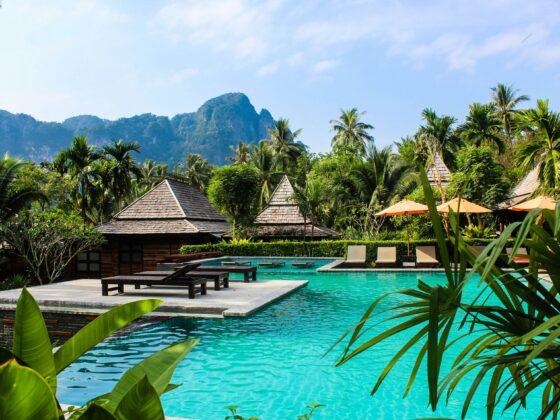 Turquoise poolside at a tropical resort with thatched villas, palm trees, and orange umbrellas, set against green hills and blue sky.