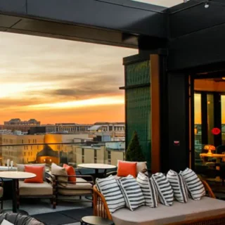 Rooftop lounge with wicker sofas and striped cushions, orange pillows, string lights, overlooking a city skyline at sunset.