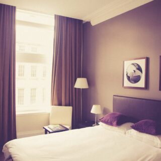 Hotel room featuring a white bed with purple pillows, two bedside lamps, a chair by a large window with heavy curtains, and circular-framed wall art on a beige wall.