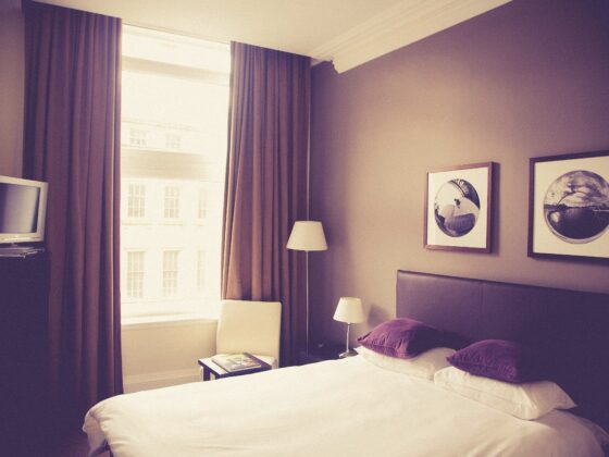 Hotel room featuring a white bed with purple pillows, two bedside lamps, a chair by a large window with heavy curtains, and circular-framed wall art on a beige wall.