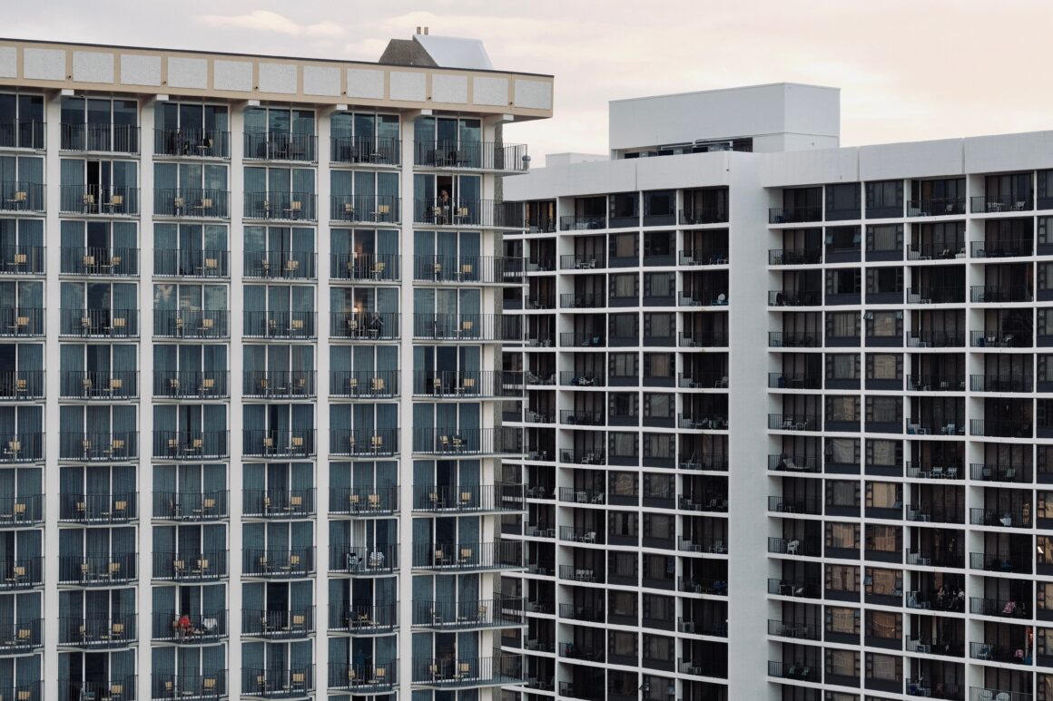 Facade of a modern apartment building with dozens of balconies and glass doors; a few residents stand on some balconies.