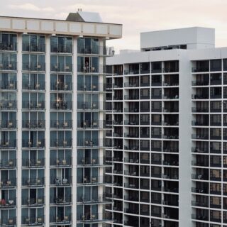 Facade of a modern apartment building with dozens of balconies and glass doors; a few residents stand on some balconies.