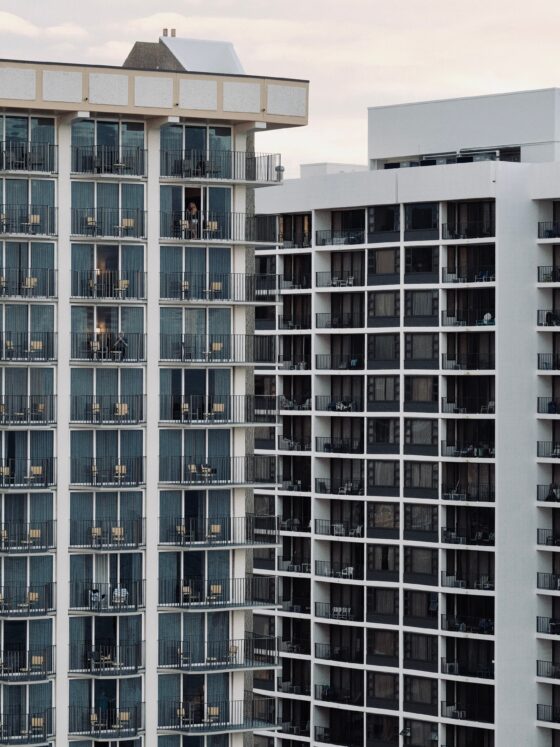 Facade of a modern apartment building with dozens of balconies and glass doors; a few residents stand on some balconies.