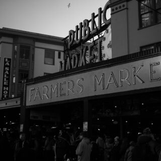 Black-and-white street scene showing a large Farmers Market sign with a clock and people gathered below near storefronts.
