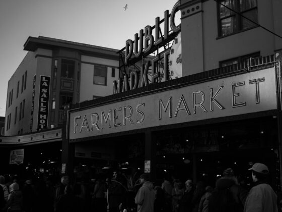 Black-and-white street scene showing a large Farmers Market sign with a clock and people gathered below near storefronts.