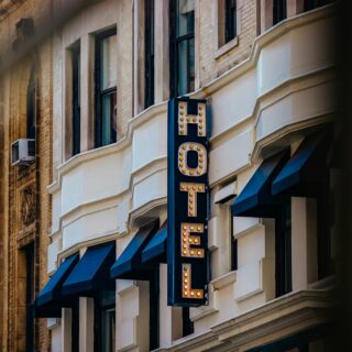 Vertical marquee sign with illuminated letters spelling 'HOTEL' on a beige brick building, with blue awnings below.