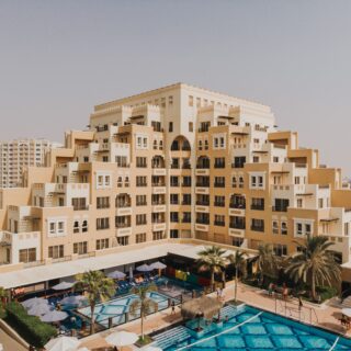 Beige multi-story resort buildings surround a bright blue pool area with lounge chairs and umbrellas on a sunny day.