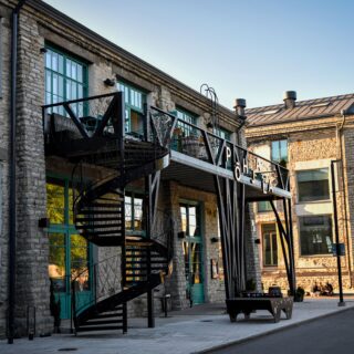 Stone building exterior with turquoise-framed windows and a black metal spiral staircase along the facade; urban plaza in foreground.