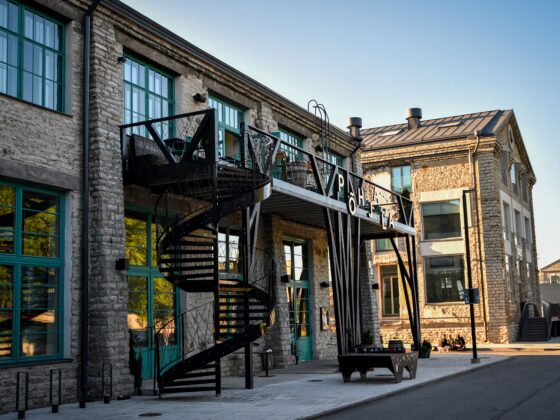 Stone building exterior with turquoise-framed windows and a black metal spiral staircase along the facade; urban plaza in foreground.