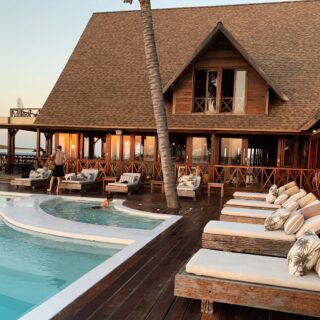 Wooden resort villa with a large slanted roof, poolside deck, and lounge chairs by an oceanfront terrace at sunset