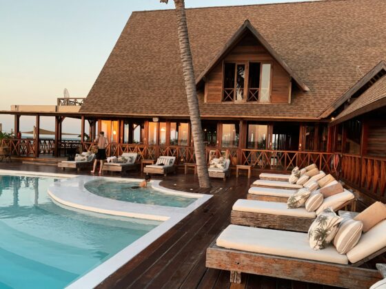 Wooden resort villa with a large slanted roof, poolside deck, and lounge chairs by an oceanfront terrace at sunset