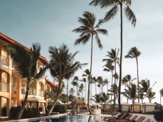 Poolside at a tropical resort: rows of wooden lounge chairs along the pool, palm trees, and orange-tan buildings in the background.