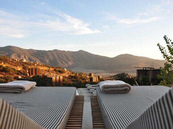 Rows of striped lounge chairs with folded towels, overlooking desert hills and mountains under a blue sky.