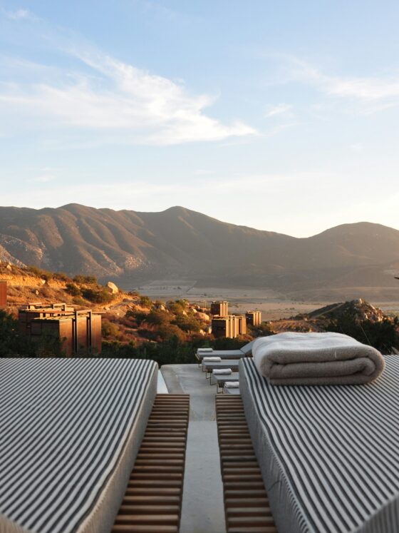 Rows of striped lounge chairs with folded towels, overlooking desert hills and mountains under a blue sky.