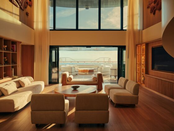 Luxurious yacht lounge with beige sofas and wooden coffee tables, glass doors open to an outdoor deck and sea view.