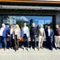 Group of ten adults in business casual clothes posing in a line outside a dark stone storefront on a sunny day.