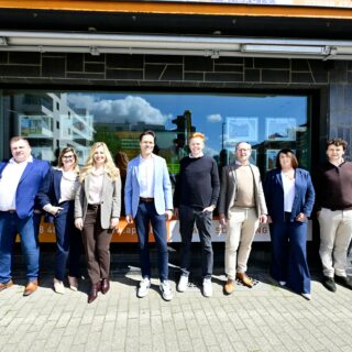 Group of ten adults in business casual clothes posing in a line outside a dark stone storefront on a sunny day.