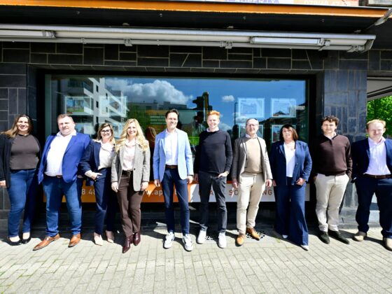 Group of ten adults in business casual clothes posing in a line outside a dark stone storefront on a sunny day.