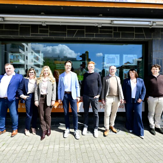 Group of ten adults in business casual clothes posing in a line outside a dark stone storefront on a sunny day.