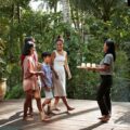 Group of five people on a wooden deck in a lush garden, as a server brings a tray of drinks intact.